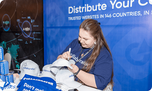 Teammitglied des Collaborator-Teams in einem gebrandeten Hemd, das Werbe-T-Shirts und Materialien an einem Stand beim SEO Mastery Summit signiert, mit den globalen Inhalteverbreitungsbotschaften von Collaborator im Hintergrund sichtbar