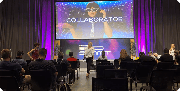Mitglieder auf einer Konferenz in Zagreb, die auf Stühlen sitzen und einem großen Bildschirm gegenüberstehen, auf dem das Promo-Video von Collaborator angezeigt wird.